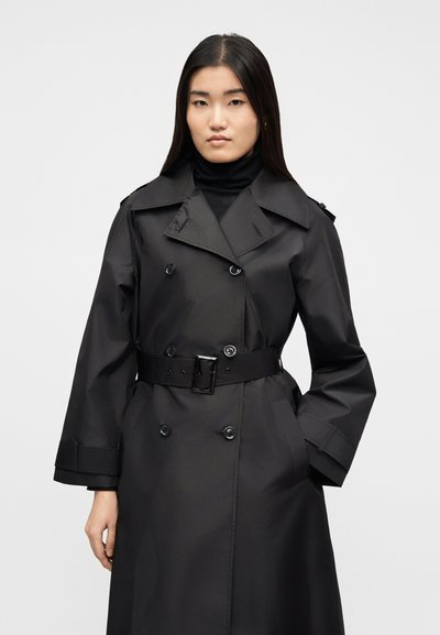 Γυναίκα που φορά μαύρο παλτό με ζώνη πάνω από μαύρο ζιβάγκο, στέκεται με το ένα χέρι στην τσέπη μπροστά σε απλό ανοιχτό φόντο.