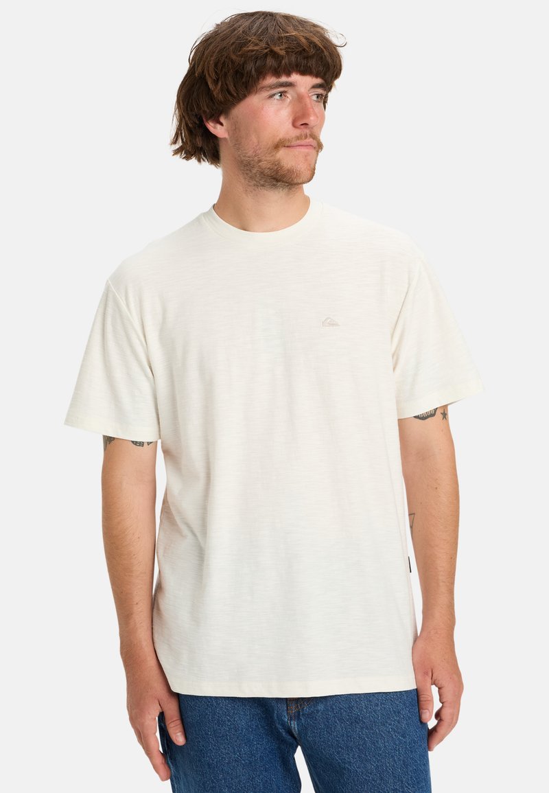 T-shirt blanc à manches courtes avec un col rond, arborant un logo subtil en gris clair sur la poitrine et un tissu texturé.