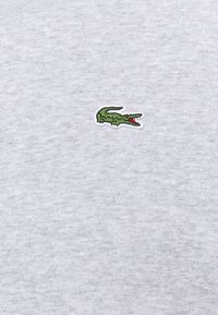 Logo de crocodile vert brodé avec bouche rouge sur fond de tissu gris clair, centré.