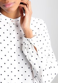 Donna che indossa una camicia bianca con pois neri, mano che tocca delicatamente il collo, labbra visibili, sfondo grigio neutro.