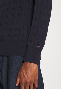 Maglione blu marino a trecce con polsini e orlo a coste. Presenta un piccolo logo rosso, bianco e blu sul maniche. Tessuto in misto cotone.