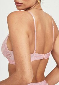 Reggiseno in pizzo rosa con spalline regolabili, schiena in rete e chiusura a gancio e occhiello, con il marchio visibile sulla banda elastica.