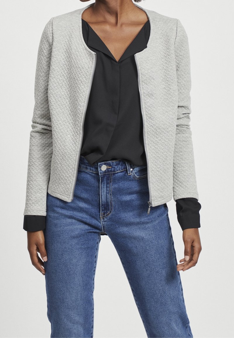 Ljusgrå texturerad zip-jacka med rund hals, med svarta manschetter och en enfärgad svart blus, parat med blå högmidjade jeans.