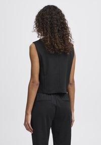 Schwarzes, kurzes, ärmelloses Oberteil mit einer geraden Rückennaht, glatter Textur und minimalistischem Design. Kombiniert mit hochgeschnittenen schwarzen Hosen mit Gesäßtaschen.