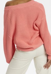 Maglione a maglia oversize con spalle scoperte, di colore rosa chiaro con motivo testurizzato. Abbinato a pantaloni color crema, che mostrano una vestibilità rilassata.