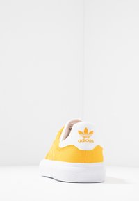 Gul canvas sneakers med vit sula och häl. Har en logotyp på baksidan och sydda detaljer längs sidorna. Rundad tåddesign.