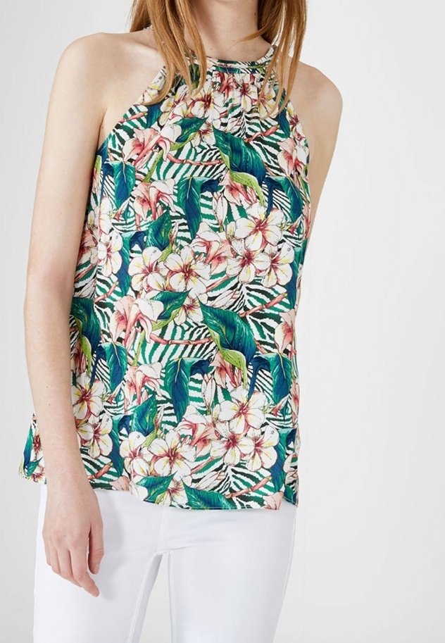 Femme portant un haut sans manches à col licou avec un motif floral tropical vert, rose et blanc, associé à un pantalon blanc.