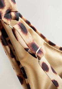 Cinturino regolabile con anello dorato su tessuto beige e marrone maculato, con dettagli in rete e cuciture.