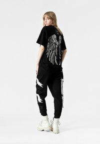 Camiseta negra con estampado de alas de ángel plateadas en la parte posterior, combinada con pantalones negros que presentan gráficos de alas en blanco y botas de combate blancas.