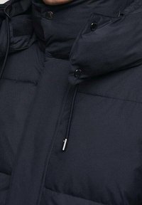 Granatowa puchowa kurtka z wysokim kołnierzem, błyszcząca tkanina, pikowane panele oraz czarne guziki. Posiada ściągacz na troczki i zapięcie na zamek błyskawiczny.