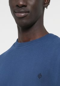 Μπλε φούτερ από μαλακό ύφασμα, με ριμπ λαιμόκοψη και μικρό, κεντητό λογότυπο στο στήθος. Casual σχεδίαση με στρογγυλή λαιμόκοψη.