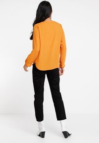 Orange long-sleeve top with a rounded neckline and a longer back, paired with black trousers and white heeled boots. Smooth fabric texture.
