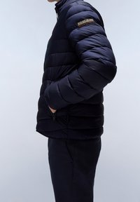 Giacca puffer blu navy con cuciture orizzontali, alto colletto, tasche con zip e logo sulla manica. Tessuto liscio e design leggero.