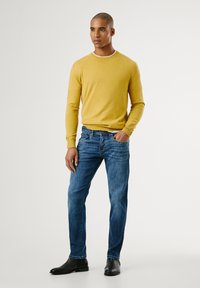 Pull jaune à col rond, jean bleu et bottes noires. Le pull a une texture lisse, un design ajusté et des poignets côtelés. Les jeans sont à coupe fuselée.