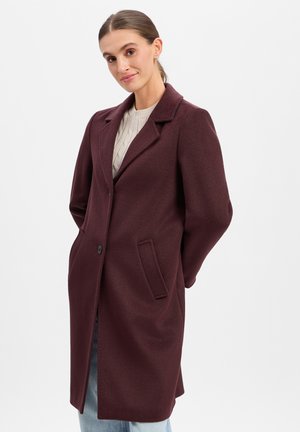 Woman wearing a long dark burgundy coat over a light knit sweater and blue jeans, standing with hands behind her back against a white background.