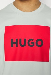 Maglione grigio chiaro con una grande grafica rettangolare rossa e il testo nero "HUGO" centrato all'interno. Tessuto morbido.