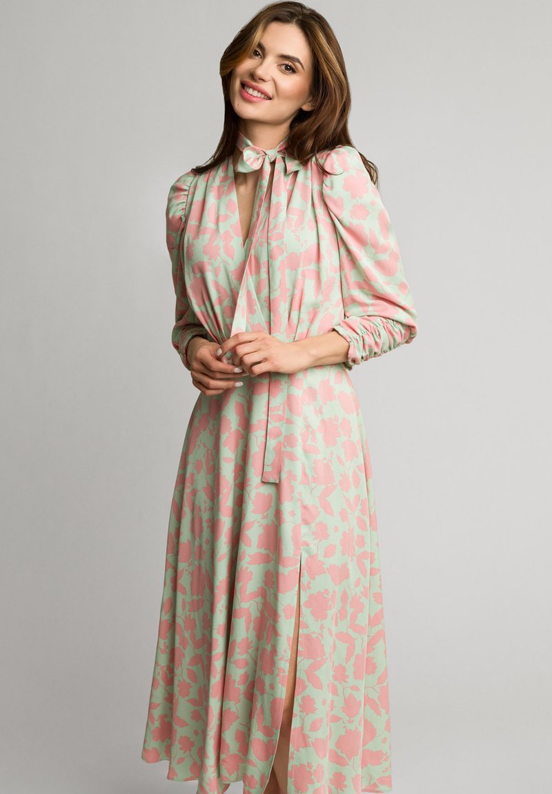 Kwiatowa sukienka z pastelowozielonym tłem, różowymi wzorami kwiatowymi, dekoltem w kształcie kokardy oraz długimi rękawami z marszczonymi mankietami.