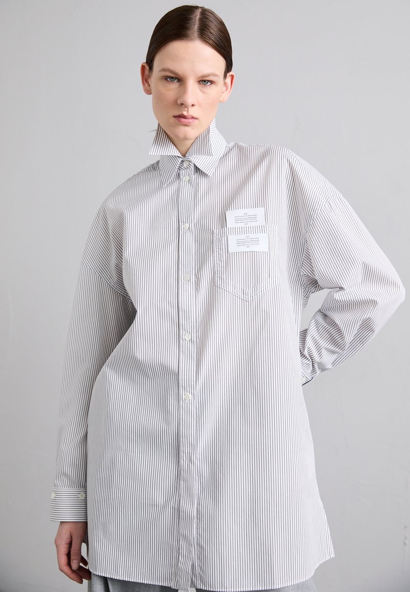 Chemise à rayures blanche et grise, dotée d'un grand col, d'une coupe ample et de deux poches poitrine avec étiquettes. Texture en coton lisse.