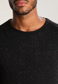 Czarna koszulka z krótkim rękawem w wzór w sploty, okrągły dekolt oraz kieszeń po lewej stronie, wykonana z miękkiego materiału.