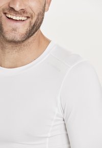 T-shirt de sport blanc à manches longues fabriqué en tissu lisse et extensible, avec une coupe ajustée et des accents en maille subtils sur les côtés.