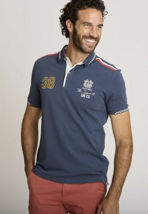 Homme aux cheveux bouclés souriant, portant un polo bleu marine avec des motifs de course et un pantalon rouge, les mains dans les poches, regardant vers la droite.