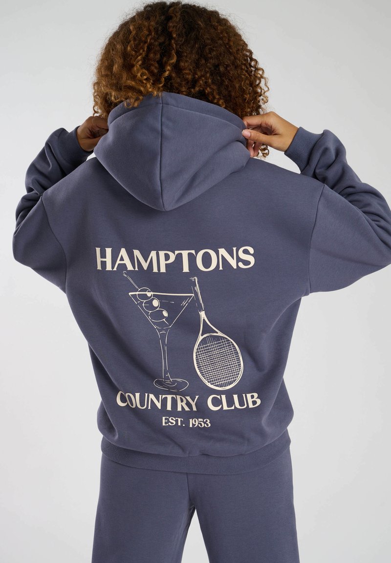 Mörkblå huvtröja med en stor gräddfärgad grafik som visar ett cocktailglas och en tennisracket, texten "HAMPTONS COUNTRY CLUB EST. 1933". Mjuk textur.