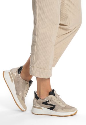 Person trägt beige Cordhosen und beige Turnschuhe mit schwarzen Akzenten und weißen Gummisohlen und steht auf einer weißen Oberfläche.