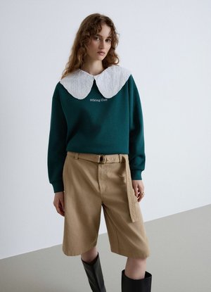 Giovane donna che indossa una felpa verde scuro con testo "Hiking Club" e grande colletto in pizzo bianco, pantaloncini beige con cintura e stivali neri al ginocchio.