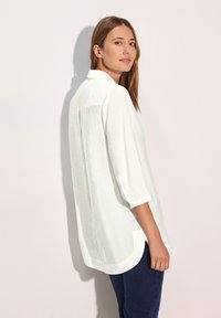 Camisa blanca de botón con cuello y mangas de tres cuartos. Presenta un dobladillo curvado y una tela ligera y suave. Se lleva con jeans oscuros.