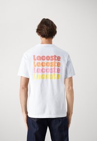 Bílá bavlněná tričko s krátkým rukávem a kulatým výstřihem, na zádech s multibarevným logem "Lacoste" v růžové, oranžové a žluté barvě.