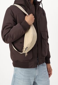 Beżowa torebka na ramię wykonana z miękkiego materiału, o zaokrąglonym kształcie, z zapięciem na suwak i regulowanym paskiem, noszona na ciemnej kurtce.