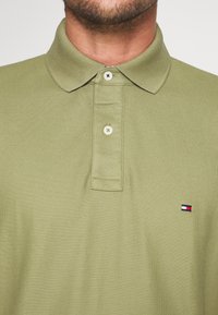 Homme portant un polo vert clair avec deux boutons blancs et un petit logo rectangulaire rouge, blanc et bleu sur la poitrine.