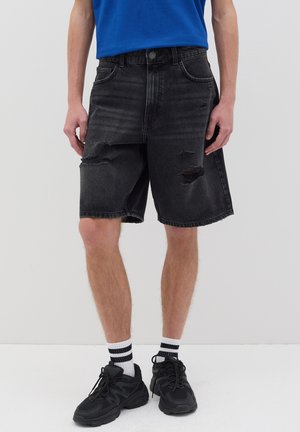 Pantalones cortos de mezclilla negra con un acabado desgastado y parches deshilachados, con un corte recto y pasadores para cinturón. Llevados con zapatillas negras y calcetines blancos.