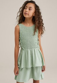 Šviesiai žalia suknelė su susmulkintu bodu, be rankovių dizainu, sluoksniuotu sijonu ir subtiliu tekstūriniu raštu. Pabrėžta šoninėmis juostelėmis.