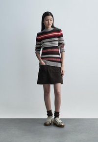Pull en maille rayé rouge, gris et noir, associé à une jupe en daim marron. Le modèle porte des baskets beiges avec des accents métalliques.