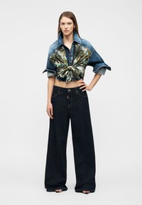 Γυναίκα που φοράει κοντή τζιν ζακέτα με τροπικό σχέδιο δεμένη στη μέση και φαρδιά τζιν παντελόνια σε σκούρο χρώμα, στέκεται με το ένα χέρι στη μέση.