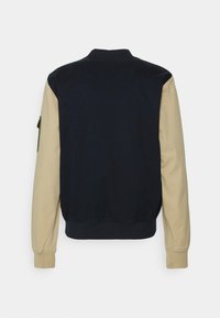 Bomberjacka med en marinblå kropp och beigefärgade ärmar, ribbade ärmslut och krage, mjukt tyg med en slät textur och med en fickdetalj på sidan.