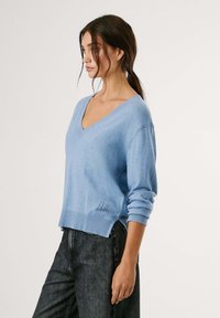 Šviesiai mėlynas, V formos megztinis, pagamintas iš minkštos mezginio medžiagos, su ilgomis rankovėmis ir banguotu apvadu. Pusėje yra mažas išsiuvinėtas detalė.