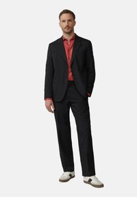 Zwarte kostuum met een gestructureerde blazer en bijpassende broek, gecombineerd met een rood shirt. Schoeisel bestaat uit witte sneakers met zwarte accenten.