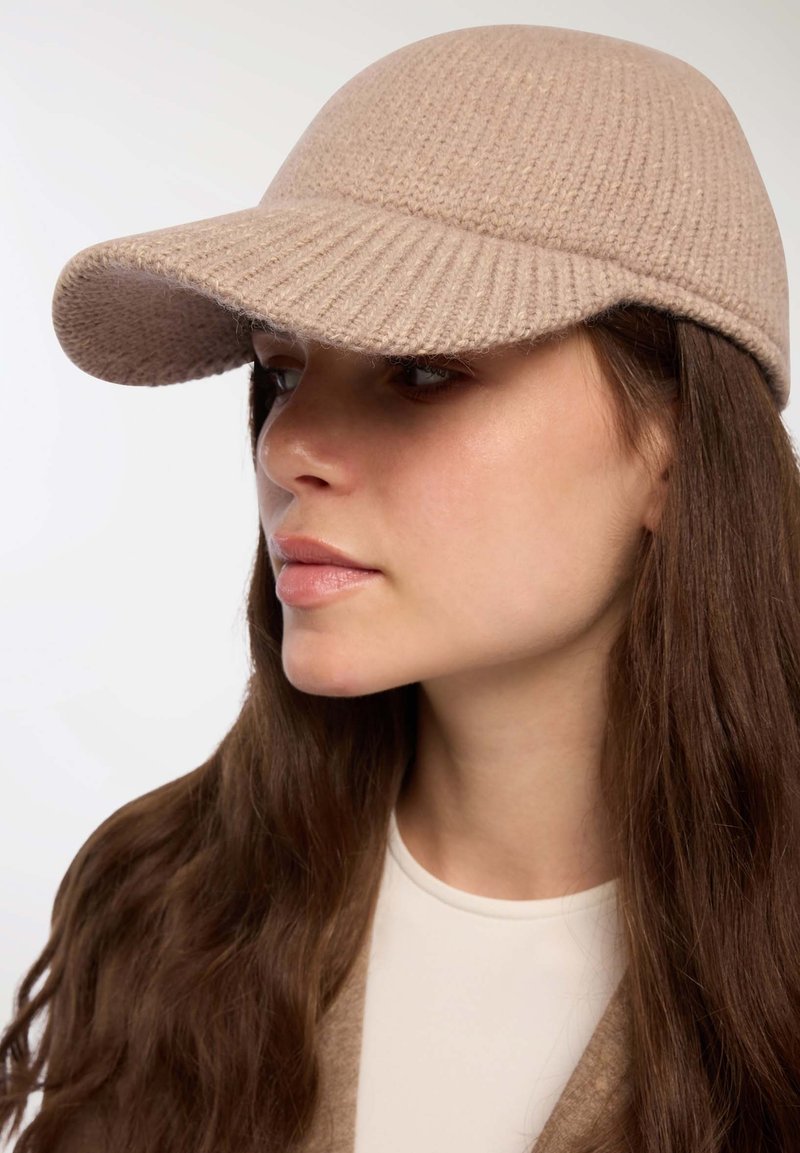Cappello di maglia beige con visiera curva, caratterizzato da una texture a coste. Il design è senza cuciture e aderisce bene alla testa.