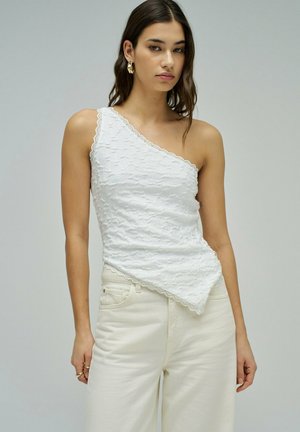 Femme portant un top blanc texturé à une épaule avec des bords festonnés et un pantalon blanc taille haute sur un fond uni.