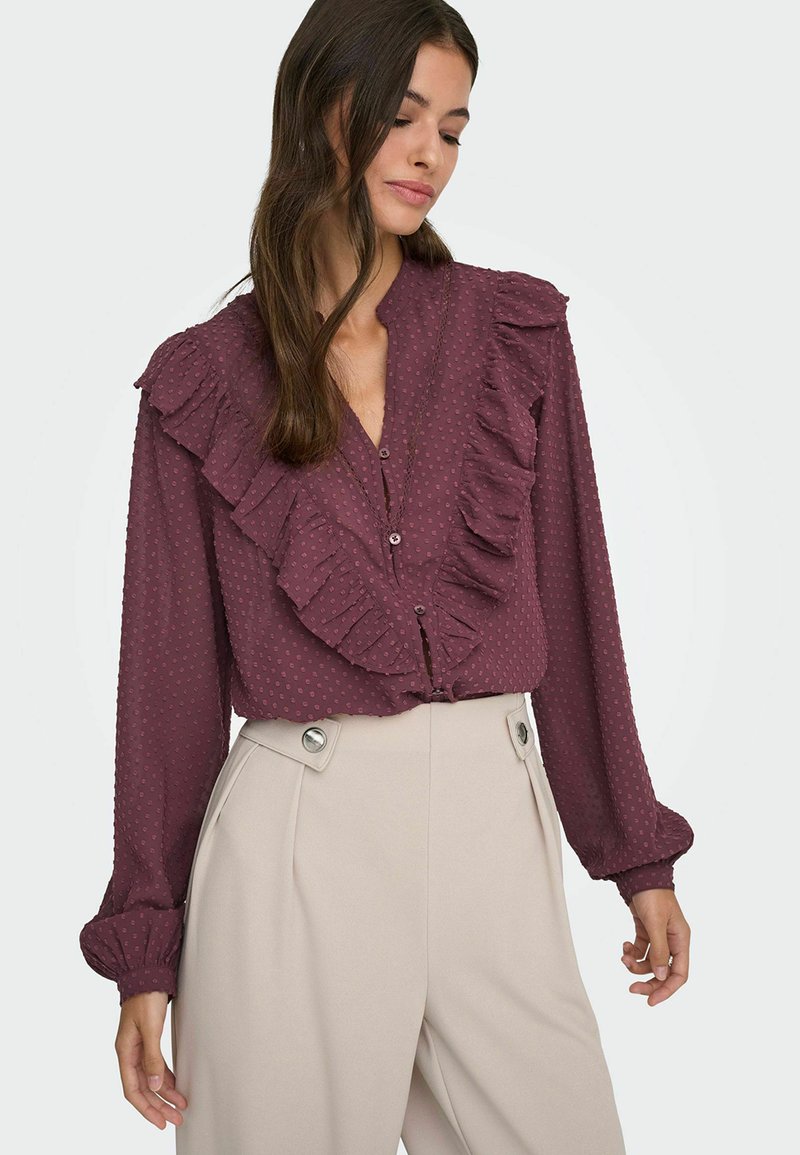 Blouse bordeaux avec devant froncé et texture à pois. Dotée d'un col et de manches longues avec des poignets bouffants.