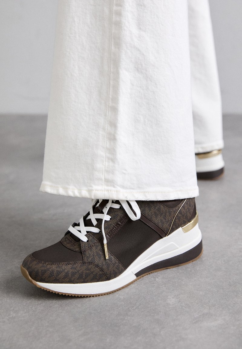 Zapatilla deportiva marrón y dorada con malla y tejido texturizado, cordones blancos y una suela blanca gruesa, parcialmente oculta por pantalones blancos.
