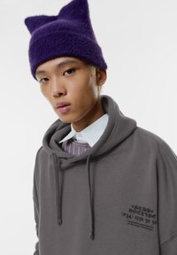Szary hoodie z dużą kieszenią z przodu i sznurkiem; posiada graficzny nadruk na klatce piersiowej. W połączeniu z fioletową, futrzaną czapką z uszami kota.