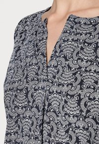 Blusa azul marino con un patrón de paisley blanco, con escote en V y tela texturizada. Diseño detallado e intrincado en todo el material.
