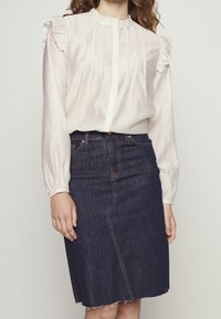 Blusa bianca con volant e bottoni sul davanti, con maniche lunghe, abbinata a una gonna di jeans blu scuro con orlo grezzo e dettaglio della cucitura sul davanti.