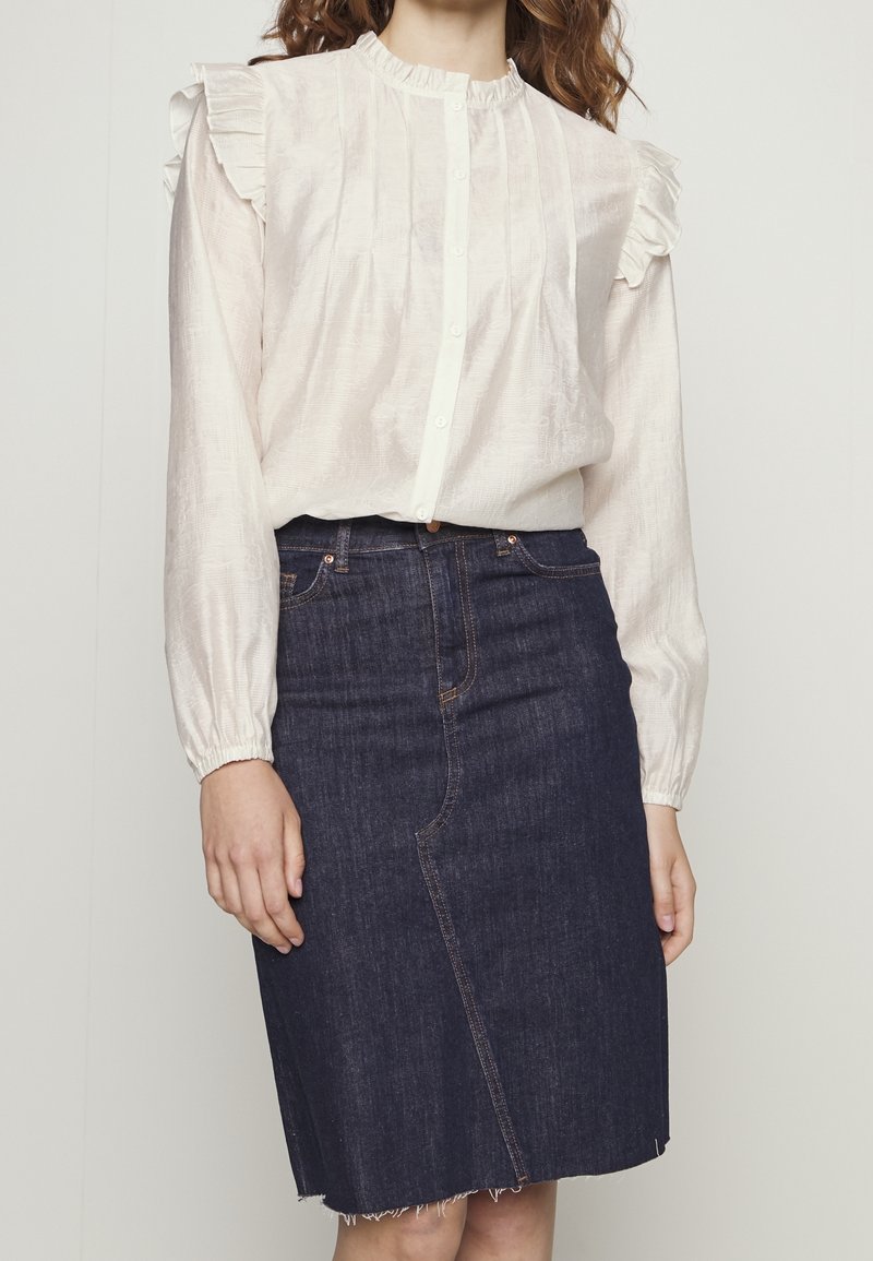 Blusa bianca con volant e bottoni sul davanti, con maniche lunghe, abbinata a una gonna di jeans blu scuro con orlo grezzo e dettaglio della cucitura sul davanti.