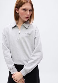 Γκρι φούτερ με φερμουάρ και γιακά, με κεντημένο λογότυπο Calvin Klein σε μαύρο. Μαλακό ύφασμα, άνετη εφαρμογή και μακριά μανίκια.