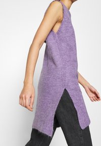 Lila ärmelloses Strickoberteil mit mittellanger Schnittführung, seitlichen Schlitzdetails und einer weichen, strukturierten Oberfläche. Wird über einer dunklen Jeanshose getragen.