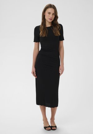 Tânără care poartă o rochie neagră midi, mulată, cu mâneci scurte și pantofi negri cu degetele descoperite, stând în picioare pe un fundal simplu, deschis la culoare.